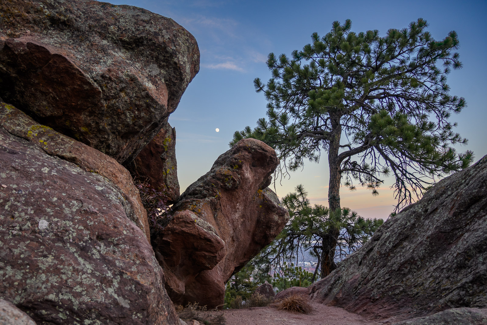 Huge red rocks covered with moss lean precariously on the edge of a mountain while the moon rises over Boulder, Colorado.
