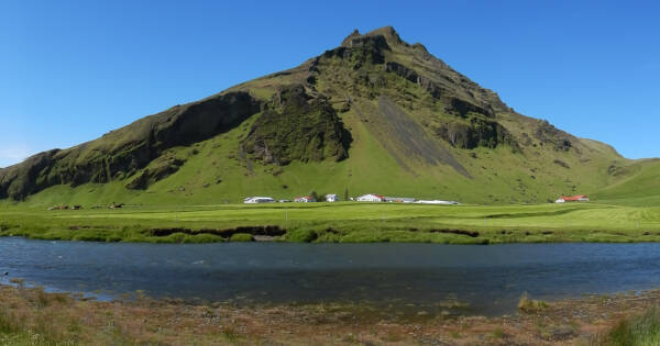 The sharp rocky peak of DrangshlÃ­Ã°ardalur protrudes from green grassy fields across the river from SkÃ³gafoss in Iceland.
