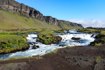 A small waterfall on the FossÃ¡lar river in South Iceland flows between green cliffs and mud.