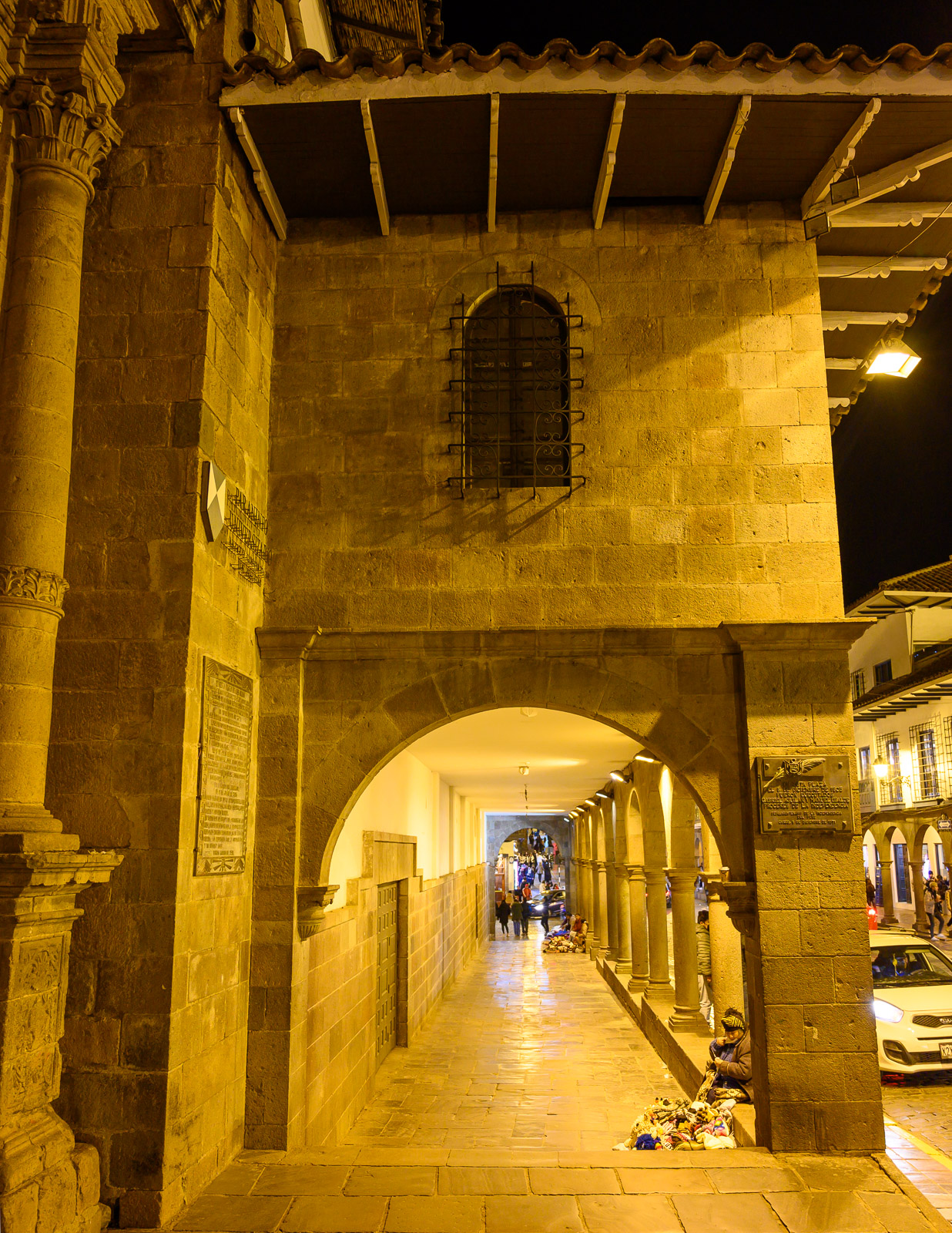 A promenade of stone arches stretches into the distance under the golden light of Plaza de Armas at night.