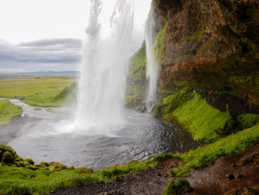 Standing in thick brown mud behind Seljalandsfoss waterfall in Iceland watching a huge stream of water drop into a circular pool surrounded by bright green vegetation.