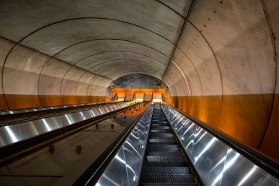 Shiny escalator descends into a concrete tunnel with an eerie orange glow.